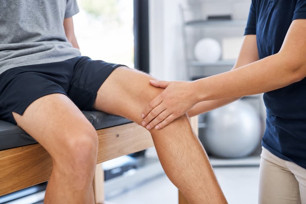 Clinical photograph showing a clinician placing their hand over a patient's patella (kneecap) to assess for clicking, popping, or grinding sensations (joint crepitus).